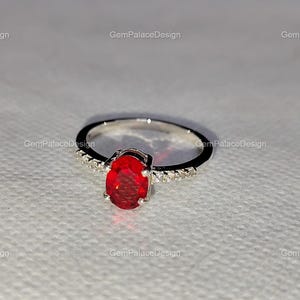 Könnte beinhalten: Ein silberner Ring mit einem großen, oval geschliffenen, leuchtend roten Edelstein als Mittelpunkt. Das Band ist mit kleinen, klaren Akzentsteinen besetzt. Der Ring ist vor einem weichen, strukturierten Hintergrund platziert, mit dem Text "GemPalaceDesign".