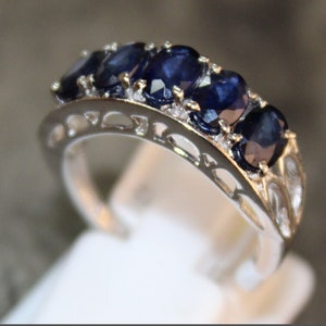 Könnte beinhalten: Ein silberner Ring mit einer Reihe ovaler blauer Saphire, die in einem filigranen, durchbrochenen Band gefasst sind.
