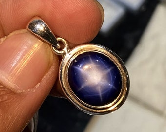 May include: A silver pendant with a blue star sapphire gemstone. The sapphire has a star-like pattern in the center.