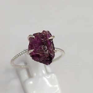 May include: A raw, uncut, dark purple gemstone ring. The gemstone is held in place by four silver prongs on a twisted silver band. The ring is displayed on a white stand against a white background.