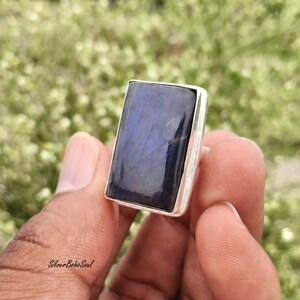 May include: A rectangular ring featuring a polished, deep blue labradorite gemstone set in a smooth silver bezel. The stone displays natural internal veining and a dark, reflective surface, securely mounted on a simple, solid silver band.