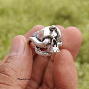 May include: A silver-toned metal ring featuring a detailed human skull design with a textured snake coiled around the cranium and jaw. The surface of the ring has an oxidized finish to highlight the intricate carved details of the bone and scales.