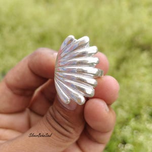 Pode incluir: Um anel de prata polida com um design em forma de leque, apresentando múltiplas cristas arredondadas e elevadas que irradiam da base. A superfície metálica reflete a luz, destacando os contornos suaves e curvos da estrutura.