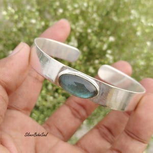 May include: A polished silver cuff bracelet featuring a smooth, oval-shaped labradorite gemstone set in the centre. The metal band has a wide, open design with a reflective surface, showcasing the iridescent blue-grey tones of the stone.