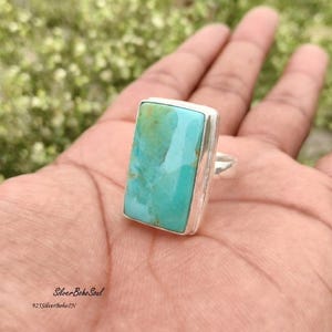 May include: A rectangular turquoise gemstone ring set in a polished silver band. The stone features a smooth surface with natural light blue and green veining. The metal band is plain and silver-toned, holding the stone in a simple bezel setting.