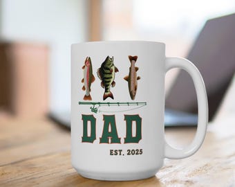 May include: White ceramic mug with illustrations of three fish above the word "DAD" in green block letters. The text "EST. 2025" is below "DAD". The mug has a curved handle and is on a wooden surface.