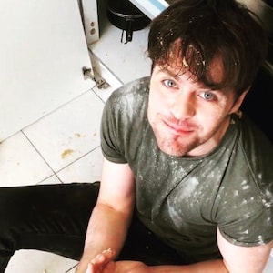 May include: A man wearing a green t-shirt covered in white powder, sitting on the floor with his legs crossed. He is looking directly at the camera and smiling.
