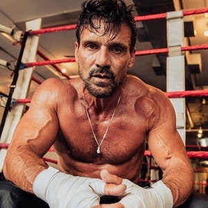 May include: A muscular man wearing boxing gloves sits in a boxing ring. He is wearing a silver necklace and has a serious expression on his face.