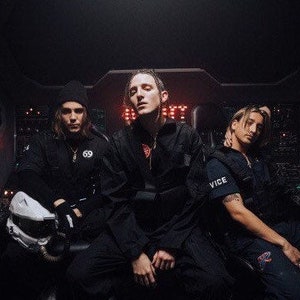 Puede incluir: Tres personas vestidas de negro posan en una habitación oscura con luces rojas. La persona del medio lleva una chaqueta negra con cremallera y un logotipo blanco. La persona de la derecha lleva un chaleco negro con la palabra "VICE" impresa.