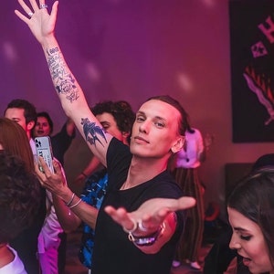 Op de afbeelding: Een man met tatoeages op zijn arm steekt zijn hand in de lucht terwijl hij naar de camera lacht. Hij draagt een zwart T-shirt en een zilveren armband. De achtergrond is een wazig beeld van mensen in een zwak verlichte kamer.