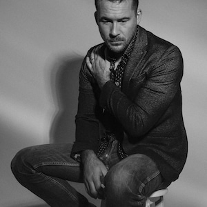 May include: A man wearing a dark grey blazer, a patterned button-down shirt, and blue jeans sits on a white stool.
