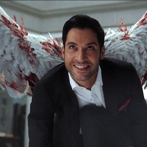 May include: A man with dark hair and a white shirt is smiling with his wings spread out behind him. The wings are white with red blood splattered on them. He is wearing a black suit jacket.