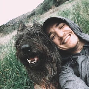 May include: A man wearing a grey hoodie and a black baseball cap smiles at the camera while holding a black shaggy dog with its tongue sticking out.