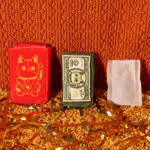 Pode incluir: Tr&ecirc;s pequenos itens de tecido coloridos s&atilde;o exibidos contra um fundo laranja texturizado. Um item retangular vermelho apresenta um design de gato dourado. Um item verde se assemelha a uma nota de d&oacute;lar com o rosto de um gato. Um item branco tamb&eacute;m est&aacute; presente.