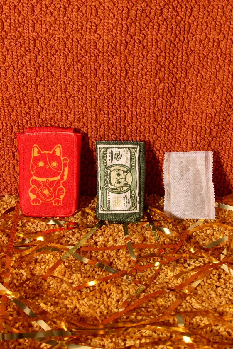 Pode incluir: Tr&ecirc;s pequenos itens de tecido coloridos s&atilde;o exibidos contra um fundo laranja texturizado. Um item retangular vermelho apresenta um design de gato dourado. Um item verde se assemelha a uma nota de d&oacute;lar com o rosto de um gato. Um item branco tamb&eacute;m est&aacute; presente.