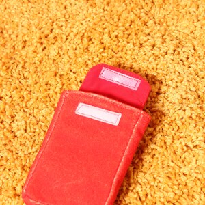 Pode incluir: Uma capa vermelha de feltro com formato retangular e uma aba presa por uma tira de velcro branca. A capa est&aacute; aberta, revelando uma etiqueta branca no interior. A capa est&aacute; sobre uma superf&iacute;cie texturizada amarelo-dourado.