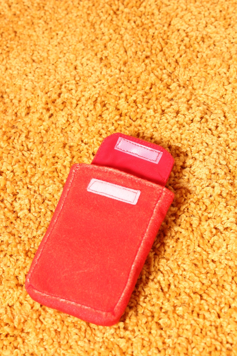 Pode incluir: Uma capa vermelha de feltro com formato retangular e uma aba presa por uma tira de velcro branca. A capa est&aacute; aberta, revelando uma etiqueta branca no interior. A capa est&aacute; sobre uma superf&iacute;cie texturizada amarelo-dourado.