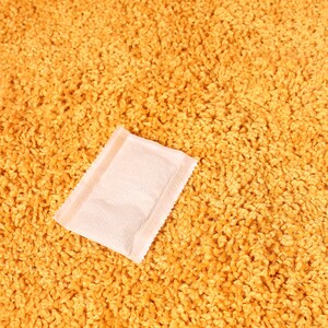 Pode incluir: Um pequeno pacote retangular rosa claro com uma superf&iacute;cie texturizada est&aacute; sobre um tapete texturizado amarelo dourado. O pacote tem uma borda selada e parece ser um item de uso &uacute;nico, possivelmente uma amostra ou um produto pequeno.