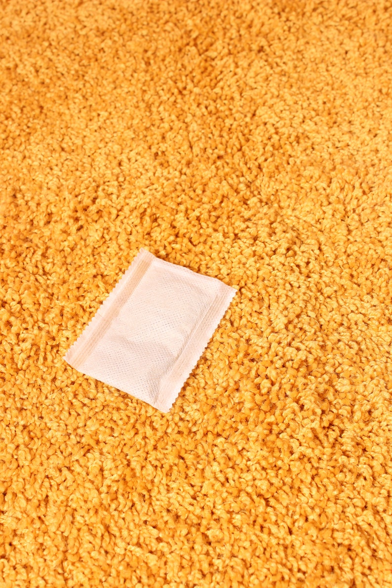 Pode incluir: Um pequeno pacote retangular rosa claro com uma superf&iacute;cie texturizada est&aacute; sobre um tapete texturizado amarelo dourado. O pacote tem uma borda selada e parece ser um item de uso &uacute;nico, possivelmente uma amostra ou um produto pequeno.