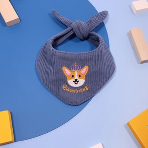 以下が含まれることがあります： 王冠をかぶったコーギーが刺繍された、青いコーデュロイのペット用バンダナ。「Queen's corgi」の文字が刺繍されています。バンダナは上部で結ばれています。背景には木製のブロックがあります。