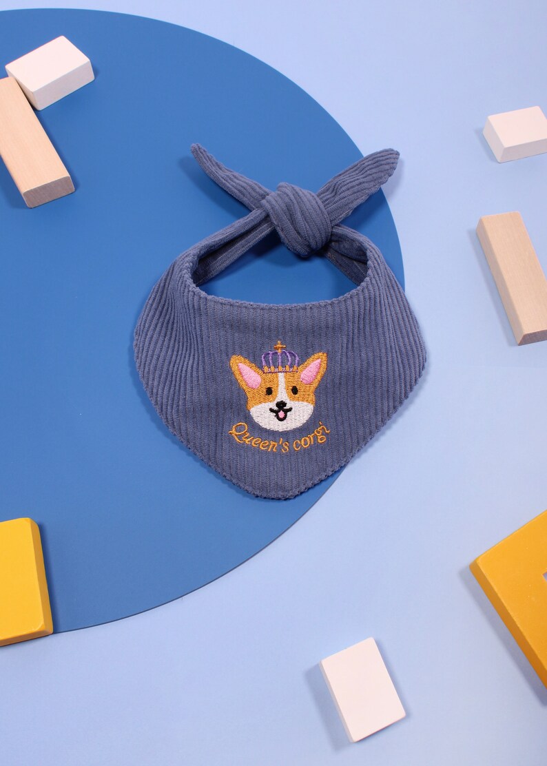 以下が含まれることがあります： 王冠をかぶったコーギーが刺繍された、青いコーデュロイのペット用バンダナ。「Queen's corgi」の文字が刺繍されています。バンダナは上部で結ばれています。背景には木製のブロックがあります。