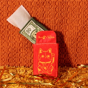 Pode incluir: Uma bolsa vermelha de gato da sorte com uma ilustra&ccedil;&atilde;o dourada, uma bolsa verde e branca com uma nota de d&oacute;lar e um pacote branco. Os itens est&atilde;o em uma superf&iacute;cie laranja texturizada com confetes dourados.