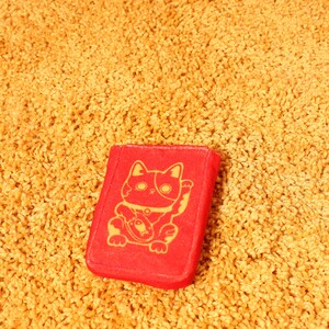 Pode incluir: Objeto retangular vermelho com uma ilustra&ccedil;&atilde;o dourada de um gato Maneki-neko, sentado e acenando com a pata. O objeto est&aacute; sobre uma superf&iacute;cie texturizada amarelo-dourado.