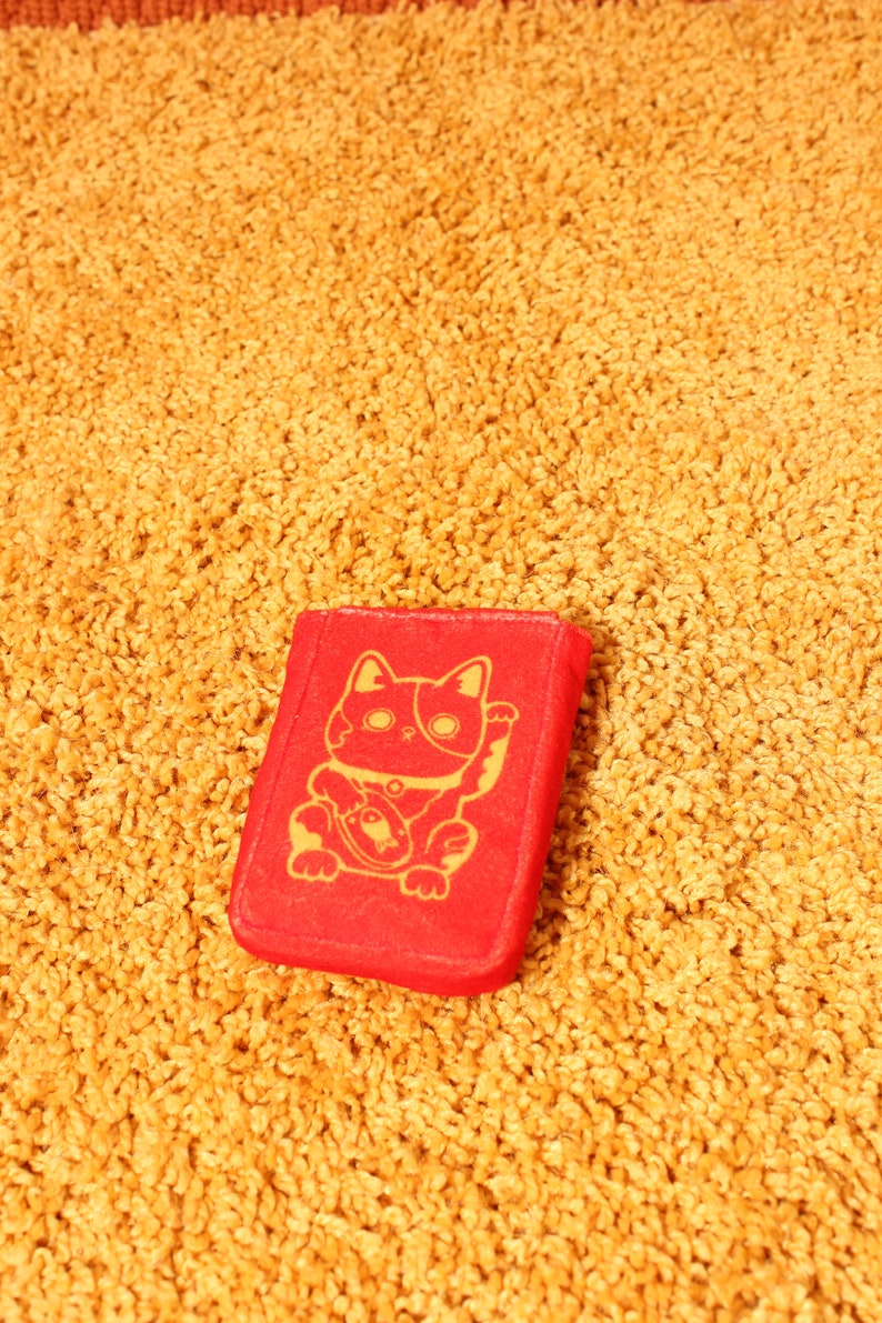 Pode incluir: Objeto retangular vermelho com uma ilustra&ccedil;&atilde;o dourada de um gato Maneki-neko, sentado e acenando com a pata. O objeto est&aacute; sobre uma superf&iacute;cie texturizada amarelo-dourado.