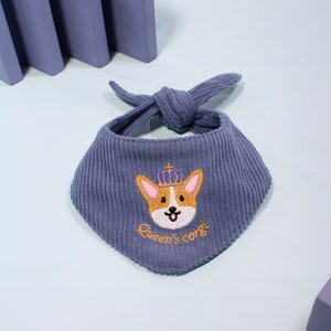 以下が含まれることがあります： 青いコーデュロイの犬用バンダナ。結び留め。王冠をかぶったコーギーの刺繍と「Queen's corgi」の文字が金色の糸で施されています。背景は水色です。