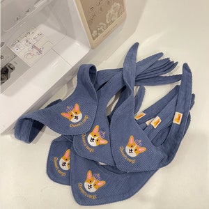 以下が含まれることがあります： 王冠をかぶったコーギーと「Queen's corgi」の文字が刺繍された、青いコーデュロイのよだれかけがいくつかあります。よだれかけはミシン近くに配置されています。