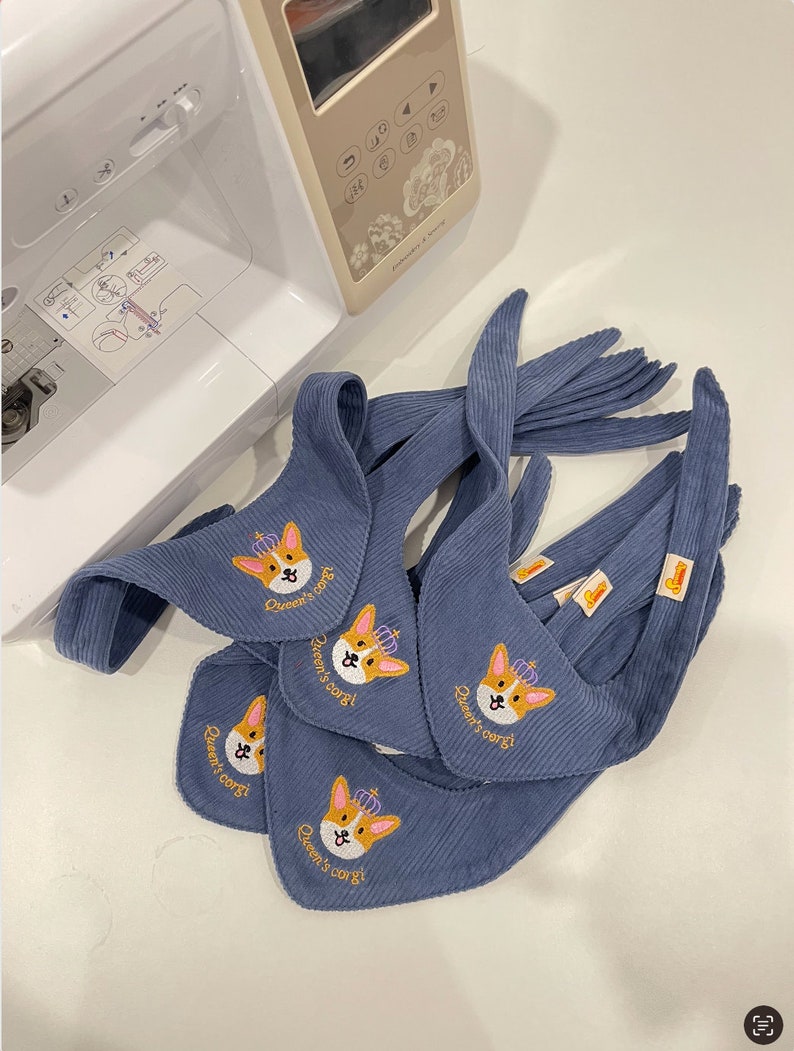 以下が含まれることがあります： 王冠をかぶったコーギーと「Queen's corgi」の文字が刺繍された、青いコーデュロイのよだれかけがいくつかあります。よだれかけはミシン近くに配置されています。