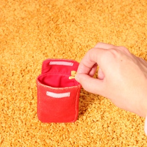 Pode incluir: Uma pequena bolsa de tecido vermelho com uma aba presa por velcro. O interior &eacute; vermelho. Uma m&atilde;o est&aacute; colocando um pequeno objeto dourado dentro da bolsa. A bolsa est&aacute; em um tapete amarelo.