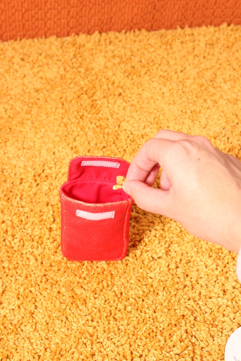 Pode incluir: Uma pequena bolsa de tecido vermelho com uma aba presa por velcro. O interior &eacute; vermelho. Uma m&atilde;o est&aacute; colocando um pequeno objeto dourado dentro da bolsa. A bolsa est&aacute; em um tapete amarelo.