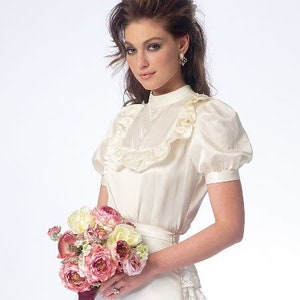 May include: A white blouse with a high ruffled collar and short sleeves. The blouse is made of a silky fabric and has a button fastening at the neck. The model is holding a bouquet of pink and white flowers.