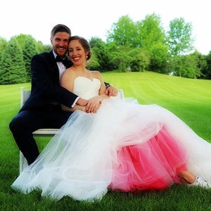 Könnte beinhalten: Eine Braut und ein Bräutigam sitzen auf einer weißen Bank in einem grasbewachsenen Feld. Die Braut trägt ein weißes Hochzeitskleid mit einem rosa Tüllrock und weißen Turnschuhen. Der Bräutigam trägt einen schwarzen Smoking.