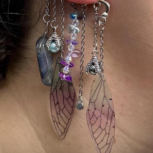 May include: A silver ear cuff with multiple dangling charms. The charms include a blue and green stone, a silver bead, a clear crystal, and two purple and clear insect wing charms.