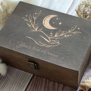 May include: A dark brown wooden box with a hinged lid and a brass latch. The lid is engraved with a crescent moon, stars, and a hand holding a branch with leaves. The text "Your text or name" is engraved below the design.