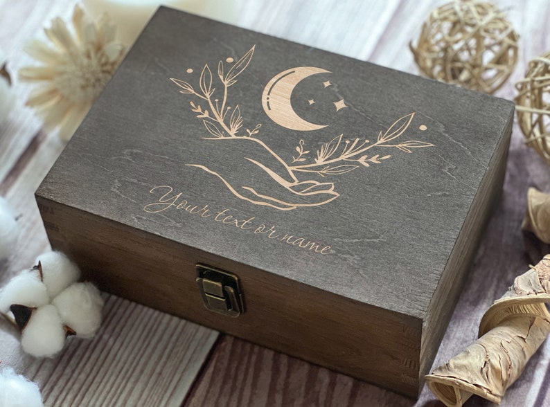May include: A dark brown wooden box with a hinged lid and a brass latch. The lid is engraved with a crescent moon, stars, and a hand holding a branch with leaves. The text "Your text or name" is engraved below the design.