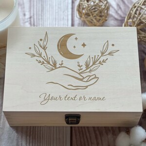 May include: A wooden box with a crescent moon and stars design, a hand holding the moon, and leafy branches. The box has a hinged lid and a brass latch. The text "Your text or name" is engraved on the front of the box.