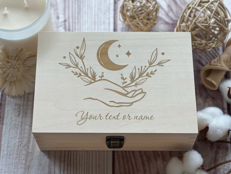 May include: A wooden box with a crescent moon and stars design, a hand holding the moon, and leafy branches. The box has a hinged lid and a brass latch. The text "Your text or name" is engraved on the front of the box.