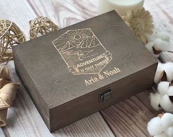 May include: A dark brown wooden box with a hinged lid and a brass latch. The box is engraved with a mountain scene and the text "Adventure is out there" and "Aria & Noah".
