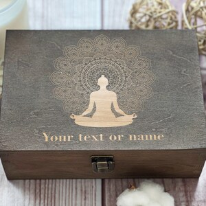 May include: A dark brown wooden box with a hinged lid and a brass latch. The box features a laser-engraved design of a person sitting in a lotus position with a mandala behind them. The text "Your text or name" is engraved below the design.