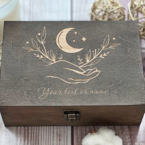 May include: A dark brown wooden box with a hinged lid and a brass latch. The top of the box is engraved with a crescent moon, stars, and two hands holding the moon. The text "Your text or name" is engraved below the design.