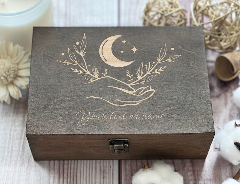 May include: A dark brown wooden box with a hinged lid and a brass latch. The top of the box is engraved with a crescent moon, stars, and two hands holding the moon. The text "Your text or name" is engraved below the design.