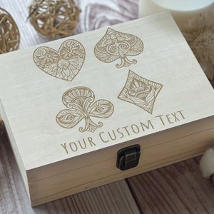 May include: A wooden box with a hinged lid, featuring an engraved design of four playing card suits: hearts, spades, diamonds, and clubs. The box has a brass latch and the text "YOUR CUSTOM TEXT" is engraved on the front.