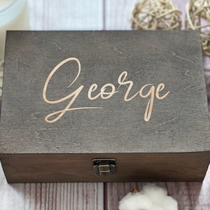 May include: A dark brown wooden box with a hinged lid and a brass latch. The box has the name "George" engraved on the top in a gold script font.