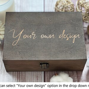 May include: A dark brown wooden box with a hinged lid and a brass latch. The box has the words "Your own design" engraved on the top in a gold color.