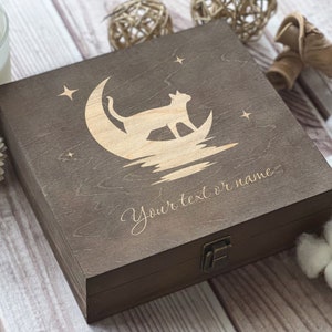 May include: A dark brown wooden box with a hinged lid and a latch. The lid is engraved with a silhouette of a cat sitting on a crescent moon with stars. The text "Your text or name" is engraved below the image.