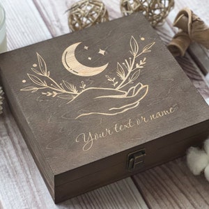 May include: A dark brown wooden box with a hinged lid. The lid is engraved with a crescent moon, stars, and a hand holding a sprig of leaves. The text "Your text or name" is engraved below the design.