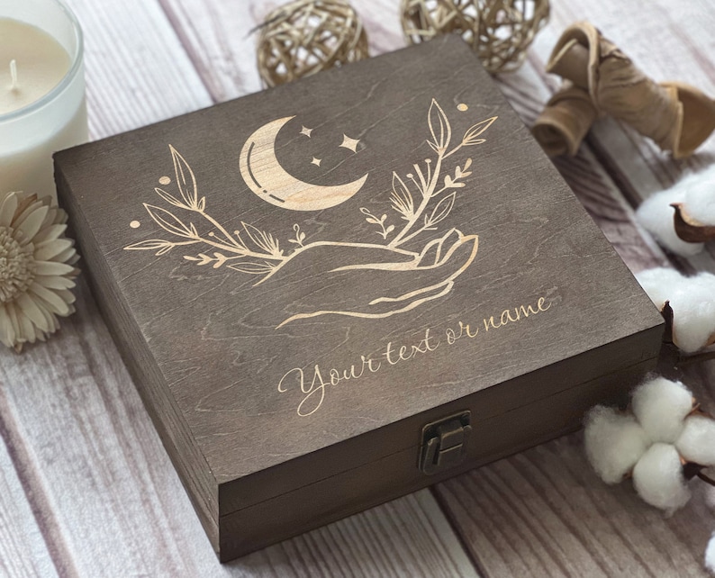 May include: A dark brown wooden box with a hinged lid. The lid is engraved with a crescent moon, stars, and a hand holding a sprig of leaves. The text "Your text or name" is engraved below the design.
